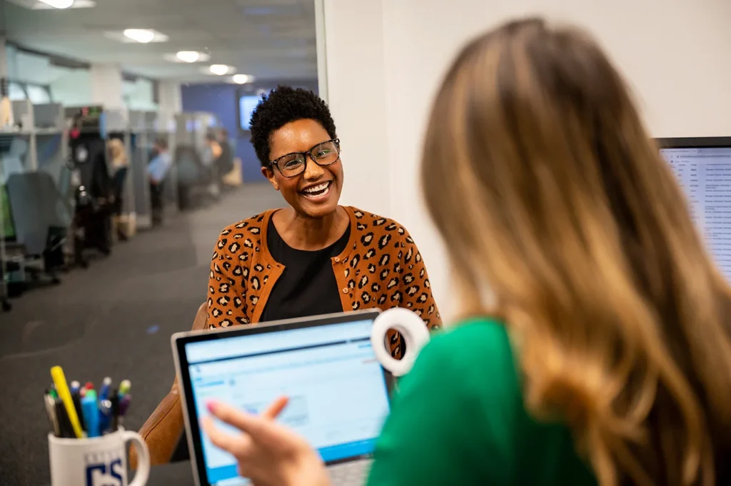 A Lady At An Office Having A Conversation With Another Lady Using A Laptop